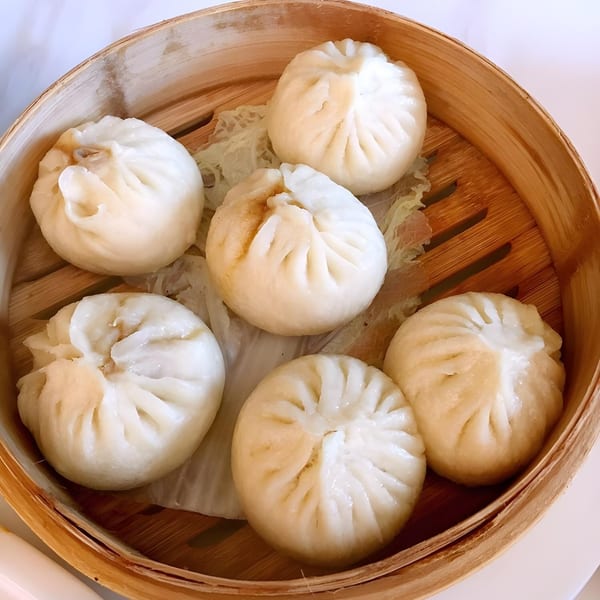 Six steamed Chinese buns, or baozi, with pleated tops, resting on lettuce in a bamboo steamer.