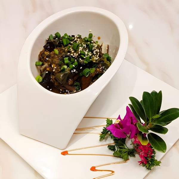 Chinese eggplant dish with green onions and sesame seeds in a white bowl, garnished with flowers.